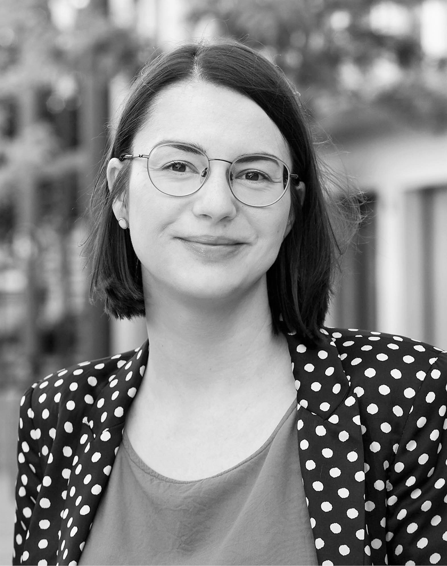 Das Bild zeigt eine Frau mit Bobfrisur, einer Brille, einem gepunkteten Blazer in schwarz-weiß.