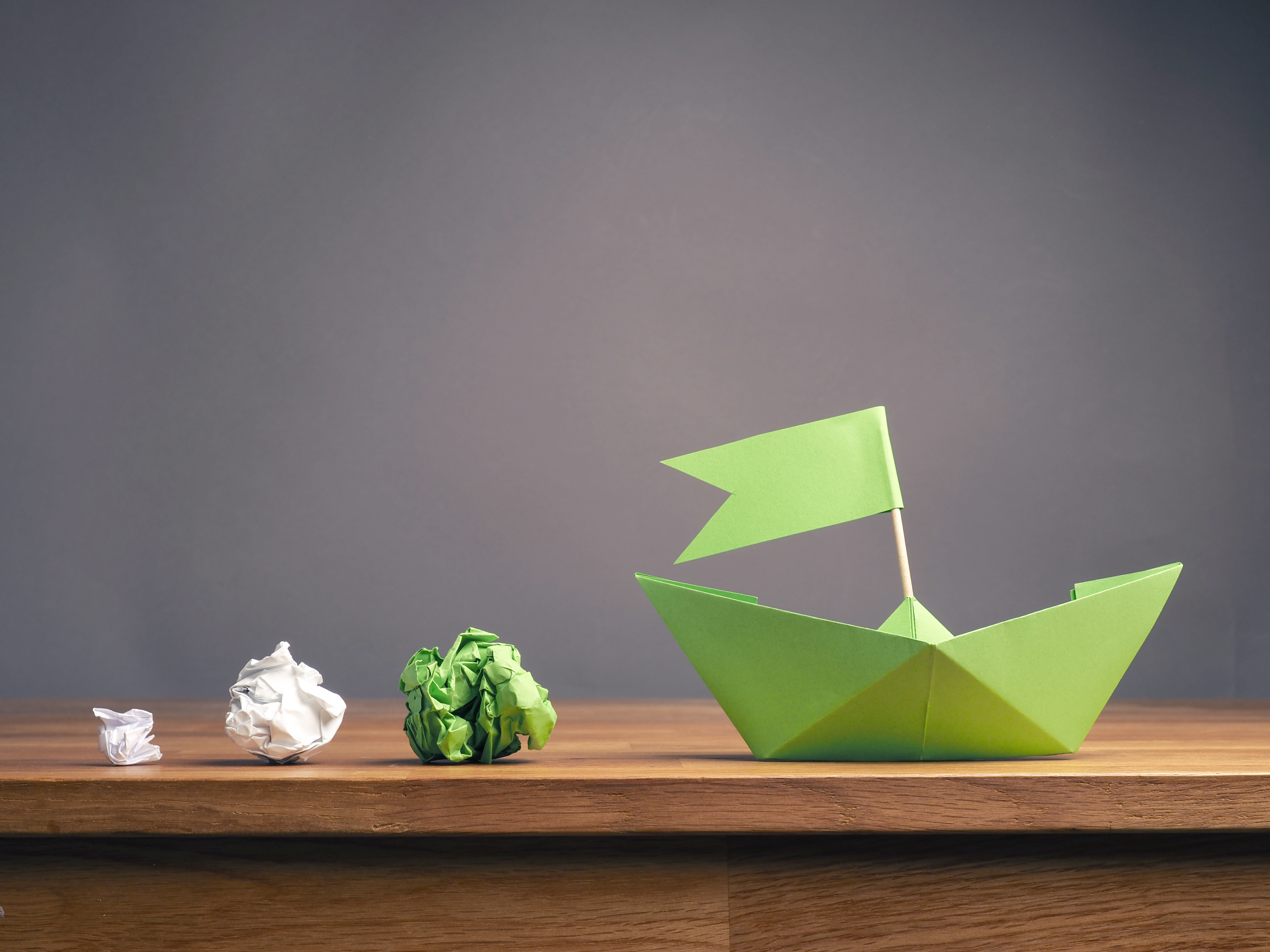 Das Bild zeigt eine Abfolge von weißen geknüllten Papierkugeln, die sich von links nach rechts zunächst grün färben und dann zu einem grünen Origamischiff werden.