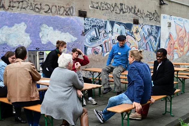 Menschen unterschiedlichen Alters sitzen draußen vor einer Graffiti-Wand auf Bierbänken und sind in einander in einem Gespräch zugewandt.