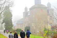 Das Bild zeigt ein Schloss im Nebel, vor dem vier Personen in herbstlicher Kleidung mit dem Rücken zur Kamera auf einem Weg stehen