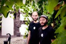 Zwei Männer in schwarzen T-Shirts vor einer bewachsenen Wand.