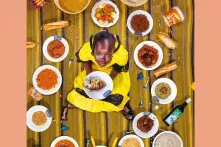 Ein Mädchen im gelben Kleid sitzt zwischen Tellern, die mit Essen gefüllt sind.