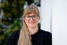 Das Bild zeigt Jennifer Korn, eine Person mit Brille, langen blonden Haaren, die lächelnd gegen eine Wand lehnt.