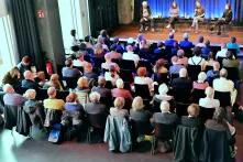 Das Bild zeigt ca. 60 Menschen von hinten, die auf Stühlen vor einer Bühne sitzen, auf der vier Personen sitzen.