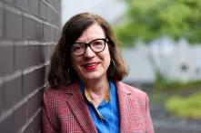 Das Bild zeigt eine Frau mit Brille und schulterlangen braunen Haaren, die in die Kamera lächelt und an einer Backsteinwand lehnt.