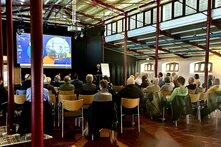 Das Bild zeigt Menschen auf Stühlen bei einer Veranstaltung, die einer Bühne zugewandt sind.