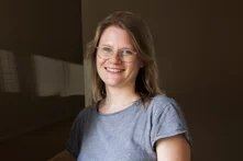 Das Bild zeigt eine mitteljunge Frau mit Brille, offenen Haaren und grauem T-Shirt, Theda Dourado. Sie lächelt in die Kamera.