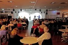Das Bild zeigt eine Aufnahme eines Publikum von hinten, welches auf Stühlen sitzt und auf die Bühne schaut. Dort stehen Personen und sprechen in Mikrofone.