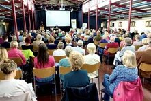 Das Bild zeigt eine Aufnahme eines Publikum von hinten, welches auf Stühlen sitzt und auf die Bühne schaut. Dort stehen Personen und sprechen in Mikrofone.