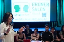 Die vortragende Person ist im Vordergrund zu sehen. Im Hintergrund sind die Zuschauenden und die Leinwand zu sehen.
