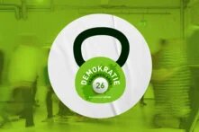 Eine grüne Kettlebell mit der Aufschrift Demokratie-Trainingslager 2026, dahinter ein verschwommenes Bild von Menschen, die sich durch einen Raum bewegen.