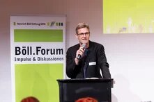 Ein Mann hält ein Mikrofon, schwarzes Sakko und Brille und spricht. Im Hintergrund ein Banner mit der Aufschrift "Böll.Forum, Impulse & Diskussionen".