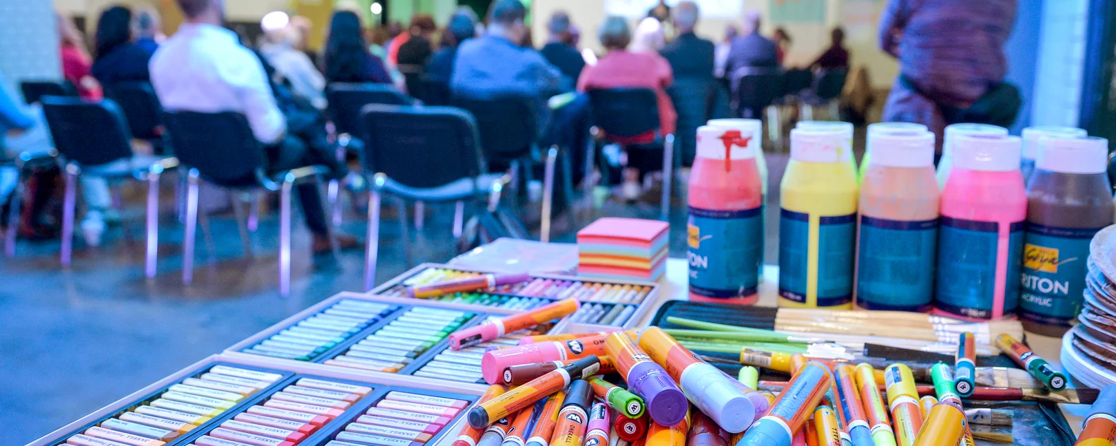 Im Vordergrund ist ein Tisch mit Stiften und Sprühflaschen (Graffiti) zu sehen, dahinter der Rücken des Publikums