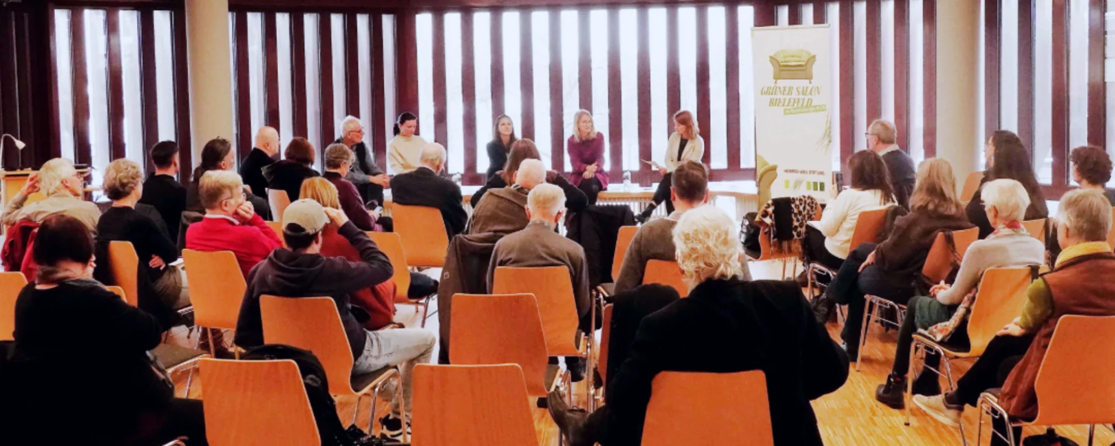 Das Bild zeigt Menschen auf Stühlen bei einer Veranstaltung.