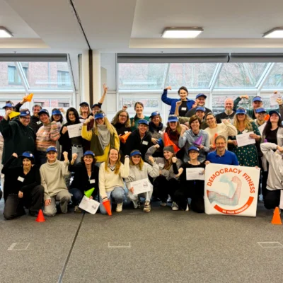40 Personen sind auf einem Gruppenbild zu sehen, einige tragen blaue Caps und Shirts, das Logo von Democracy Fitness ist auf einem Banner zu sehen.