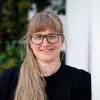 Das Bild zeigt Jennifer Korn, eine Person mit Brille, langen blonden Haaren, die lächelnd gegen eine Wand lehnt.
