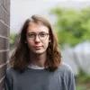 Das Bild zeigt einen jungen Mann mit Brille und schulterlangen Haaren, der gegen eine Backsteinwand lehnt.