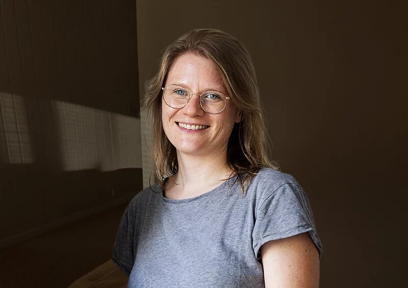 Das Bild zeigt eine mitteljunge Frau mit Brille, offenen Haaren und grauem T-Shirt, Theda Dourado. Sie lächelt in die Kamera.