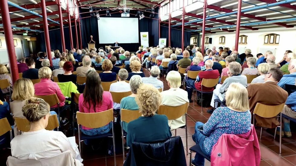 Das Bild zeigt eine Aufnahme eines Publikum von hinten, welches auf Stühlen sitzt und auf die Bühne schaut. Dort stehen Personen und sprechen in Mikrofone.