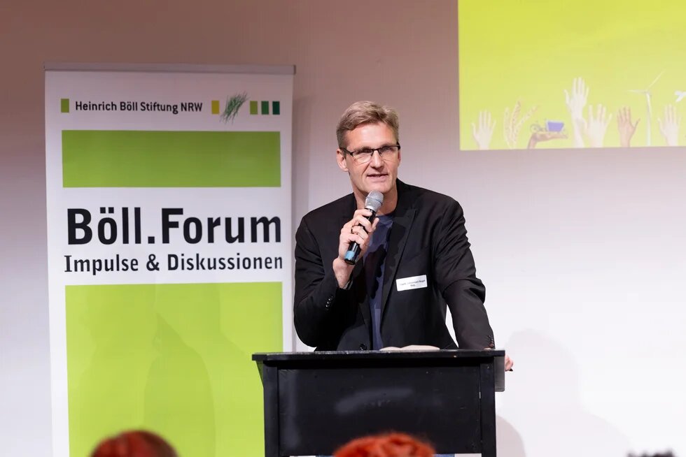 Ein Mann hält ein Mikrofon, schwarzes Sakko und Brille und spricht. Im Hintergrund ein Banner mit der Aufschrift "Böll.Forum, Impulse & Diskussionen".