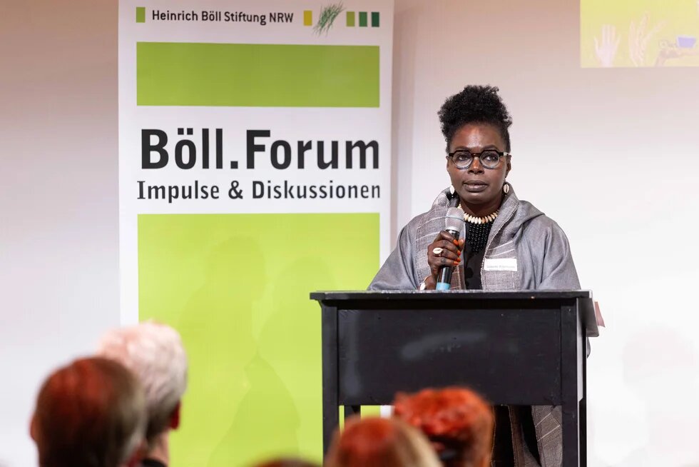 Eine schwarze Frau steht an einem Redepult, hält ein Mikrofon in der Hand, im Hintergrund ein grünes Banner mit der Aufschrift Böll-Forum: Impulse & Diskussionen.