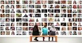 Drei Menschen sitzen auf einer Bank vor einer großen Foto-Wand mit vielen unterschiedlichen Gesichtern.